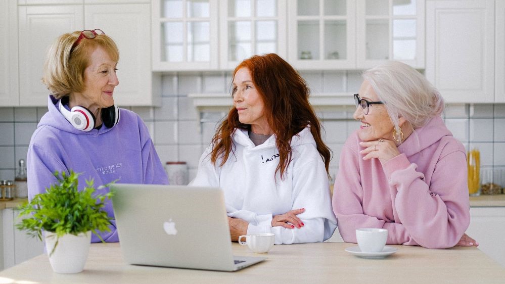 three women engage in a conversation in a kitchen with a laptop open wearing workout attire